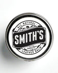 A metal tin of Smith's Leather Balm rests against a white backdrop. It features the company's black logo on a white sticker, with the words "all natural leather balm" and "made in Maine" underneath the company's name.