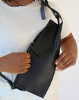 A woman wears a black leather fanny pack shoulder bag against a white background. It shows her unzipping the zipper close to her chest.