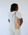 A woman wears a natural, undyed leather fanny pack shoulder bag against a white background. Her back faces the camera and the bag rests against her back.