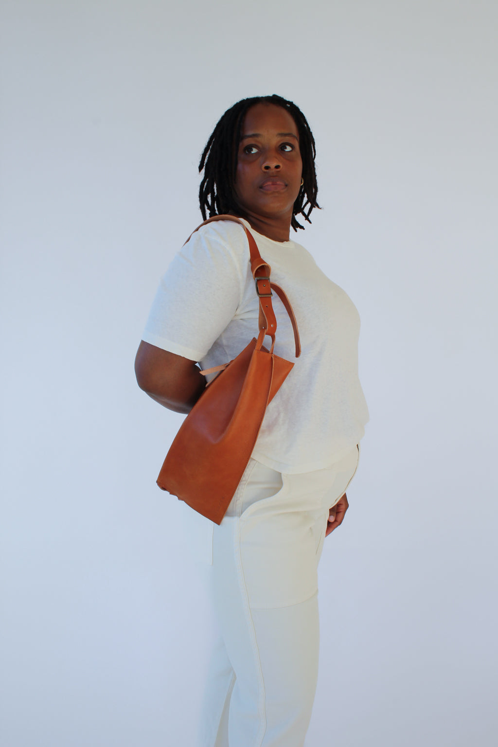 A woman wears a light brown chestnut leather fanny pack shoulder bag against a white background. She looks over her shoulder.