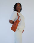 A woman wears a light brown chestnut leather fanny pack shoulder bag against a white background. She looks over her shoulder.