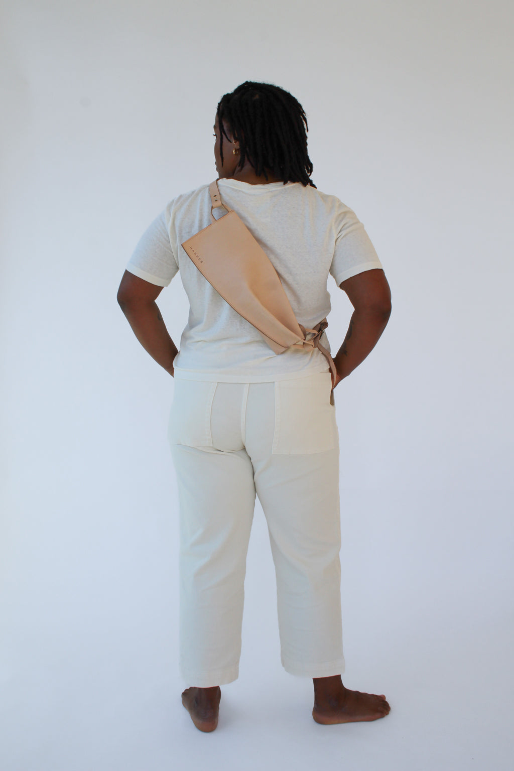 A woman wears a natural, undyed leather fanny pack shoulder bag against a white background. Her back faces the camera and the bag rests against her back.