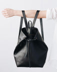 A woman holds out her arm, which is looped through the shoulder straps of a black leather backpack. The top corners of the backpack are cinched with straps that knot over a zipper, creating a unique shape.