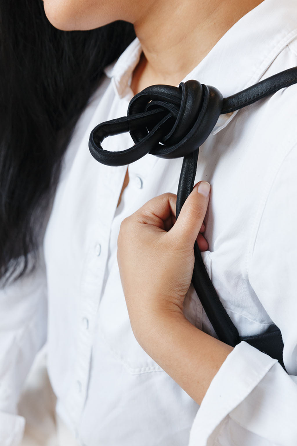 A woman sits on a chair, wearing a black leather shoulder bag over her shoulder. The shoulder strap is tied so a knot sits over her shoulder. This is a detail of the knot over her shoulder.