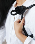 A woman sits on a chair, wearing a black leather shoulder bag over her shoulder. The shoulder strap is tied so a knot sits over her shoulder. This is a detail of the knot over her shoulder.