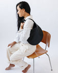 A woman sits on a chair, wearing a black leather shoulder bag over her shoulder. The shoulder strap is tied so a knot sits over her shoulder. The leather is soft and shiny, and the bag is a half moon shape.