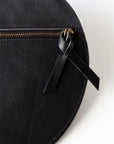 A circular black leather clutch rests against a white background with the reverse side and zipper visible. The zipper includes a matching zipper pull. The focus is on the zipper pull.