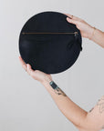 A woman's arms are visible, holding a circular black leather clutch with the reverse side and zipper visible. The zipper includes a matching zipper pull.