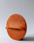A circular cognac brown leather clutch rests against a white background.