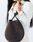 A woman holds the knotted strap of a dark brown leather shoulder bag to her chest, so that the knot is visible.