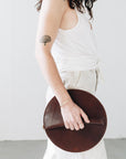 A woman holds a circular dark brown leather clutch in her hands as she walks.