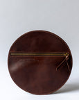 A circular dark brown leather clutch rests against a white background with the reverse side and zipper visible. The zipper includes a matching zipper pull.