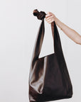 A woman's arm is visible, holding out the strap of a large, soft dark brown leather shoulder bag.