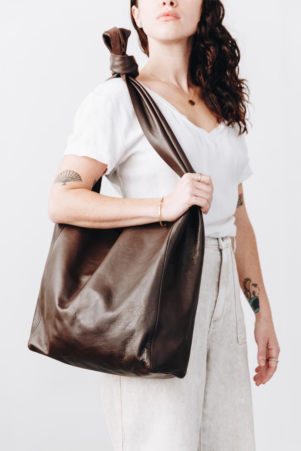 A woman wears a large, soft dark brown leather shoulder bag. The wide strap is knotted at the top where it rests on her shoulder.