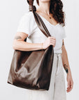 A woman wears a large, soft dark brown leather shoulder bag. The wide strap is knotted at the top where it rests on her shoulder.