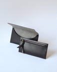A dark brown leather envelope wallet sits against a white background. It has the "Harper" logo heat stamped at the bottom, with the top flap secured by a brass button stud. The inner component is in front of the outer one, showing a zipper and matching leather pull.