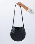A woman's arm is visible, holding out a black leather shoulder bag by the thin strap. The leather is soft and shiny, and the bag is a half moon shape.