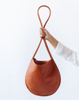 A woman's arm is visible, holding out a cognac leather shoulder bag by the thin strap. The leather is soft and shiny, and the bag is a half moon shape.