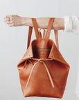 A woman holds out her arm, which is looped through the shoulder straps of a cognac brown leather backpack. The top corners of the backpack are cinched with straps that knot over a zipper, creating a unique shape.