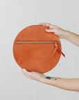 A woman's arms are visible, holding out a circular natural leather clutch with the reverse side and zipper visible. The zipper includes a matching zipper pull.