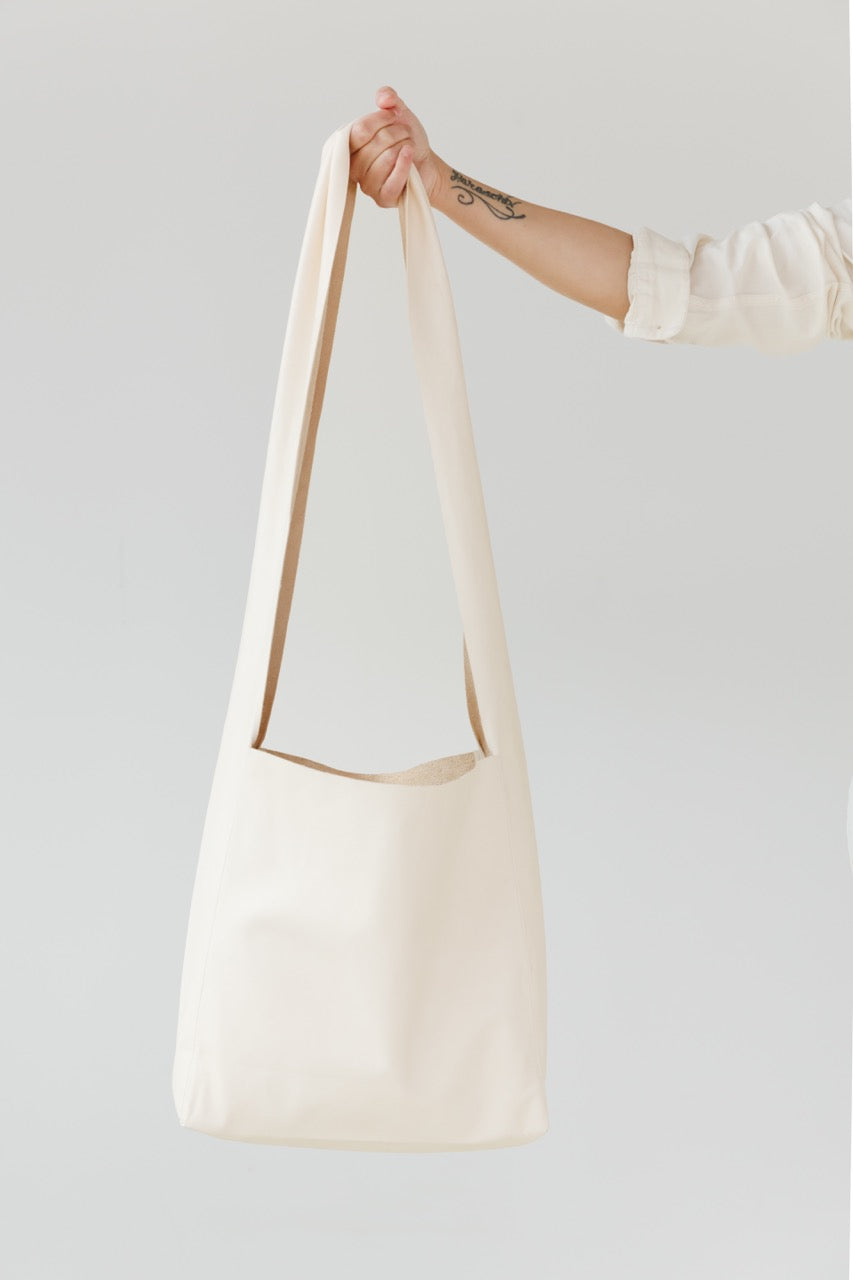 A woman's arm is visible, holding out the strap of a large, soft undyed, natural leather shoulder bag.