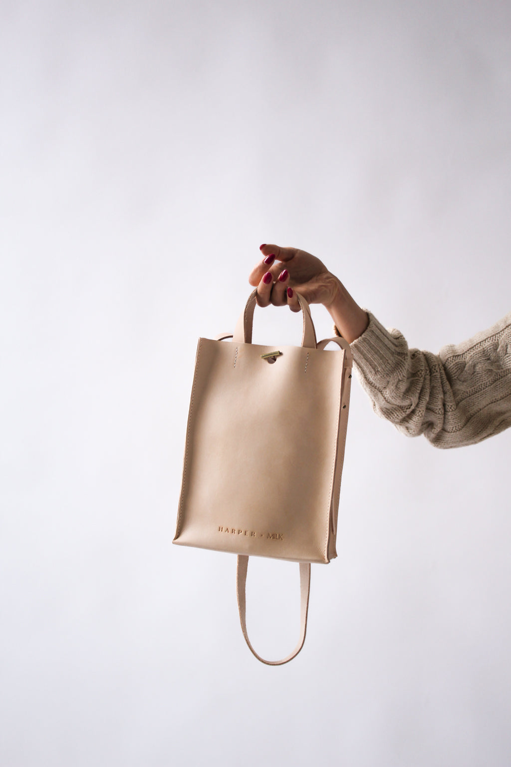 A woman's arm is visible, and she gently holds a natural undyed leather, rectangular shoulder bag in her open hand. The Harper x Milk heat stamped logo is visible on the bag.