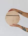 A woman's arms are visible, holding a circular natural leather clutch with the reverse side and zipper visible. The zipper includes a matching zipper pull.
