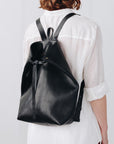 A woman faces away from the camera, wearing a black leather backpack. The top corners of the backpack are cinched with straps that knot over a zipper, creating a unique shape.