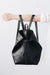 A woman holds out her arm, which is looped through the shoulder straps of a black leather backpack. The top corners of the backpack are cinched with straps that knot over a zipper, creating a unique shape.