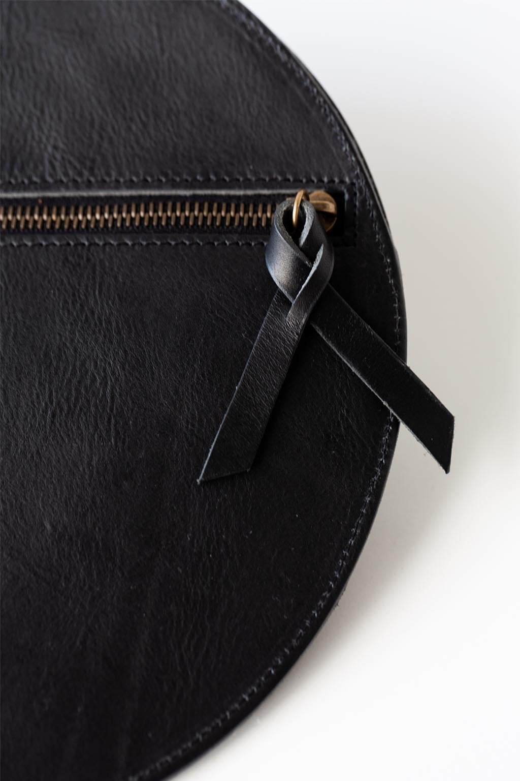 A circular black leather clutch rests against a white background with the reverse side and zipper visible. The zipper includes a matching zipper pull. The focus is on the zipper pull.