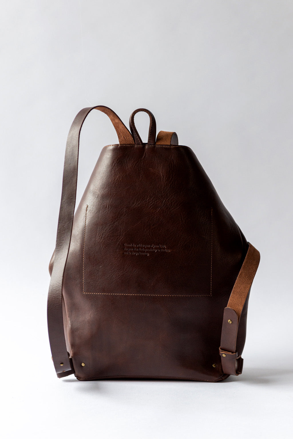 A dark brown leather backpack sits against a white backdrop, showing the reverse of the backpack with stitching for the interior pocket.