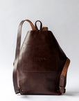 A dark brown leather backpack sits against a white backdrop, showing the reverse of the backpack with stitching for the interior pocket.