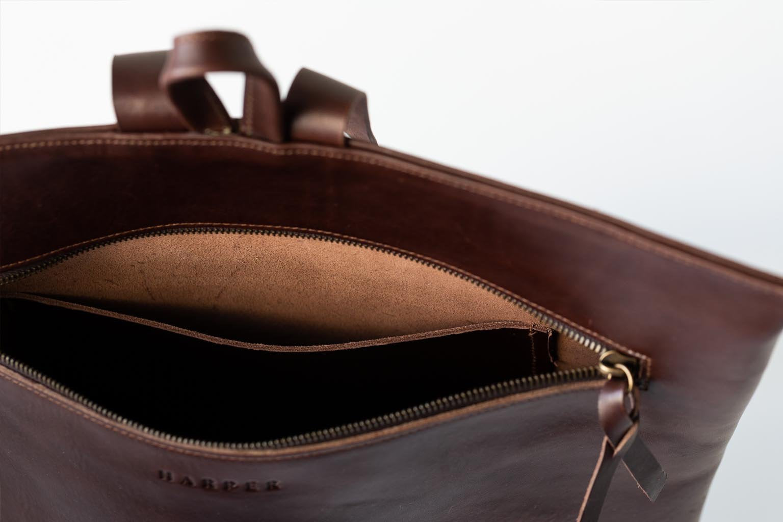 A detail of a dark brown leather backpack showing the top strap, zipper, and interior pocket of the backpack.