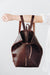 A woman holds out her arm, which is looped through the shoulder straps of a dark brown leather backpack. The top corners of the backpack are cinched with straps that knot over a zipper, creating a unique shape.