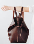A woman holds out her arm, which is looped through the shoulder straps of a dark brown leather backpack. The top corners of the backpack are cinched with straps that knot over a zipper, creating a unique shape.