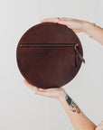 A woman's arms are visible, holding a circular dark brown leather clutch with the reverse side and zipper visible. The zipper includes a matching zipper pull.