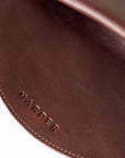 A circular dark brown leather clutch rests against a white background. The focus is on the embossed "Harper" logo and the stitching.