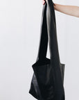 A woman's arm is visible, holding out the strap of a large, soft black leather shoulder bag.