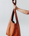 A woman's arm is visible, holding out the strap of a large, soft cognac brown leather shoulder bag.