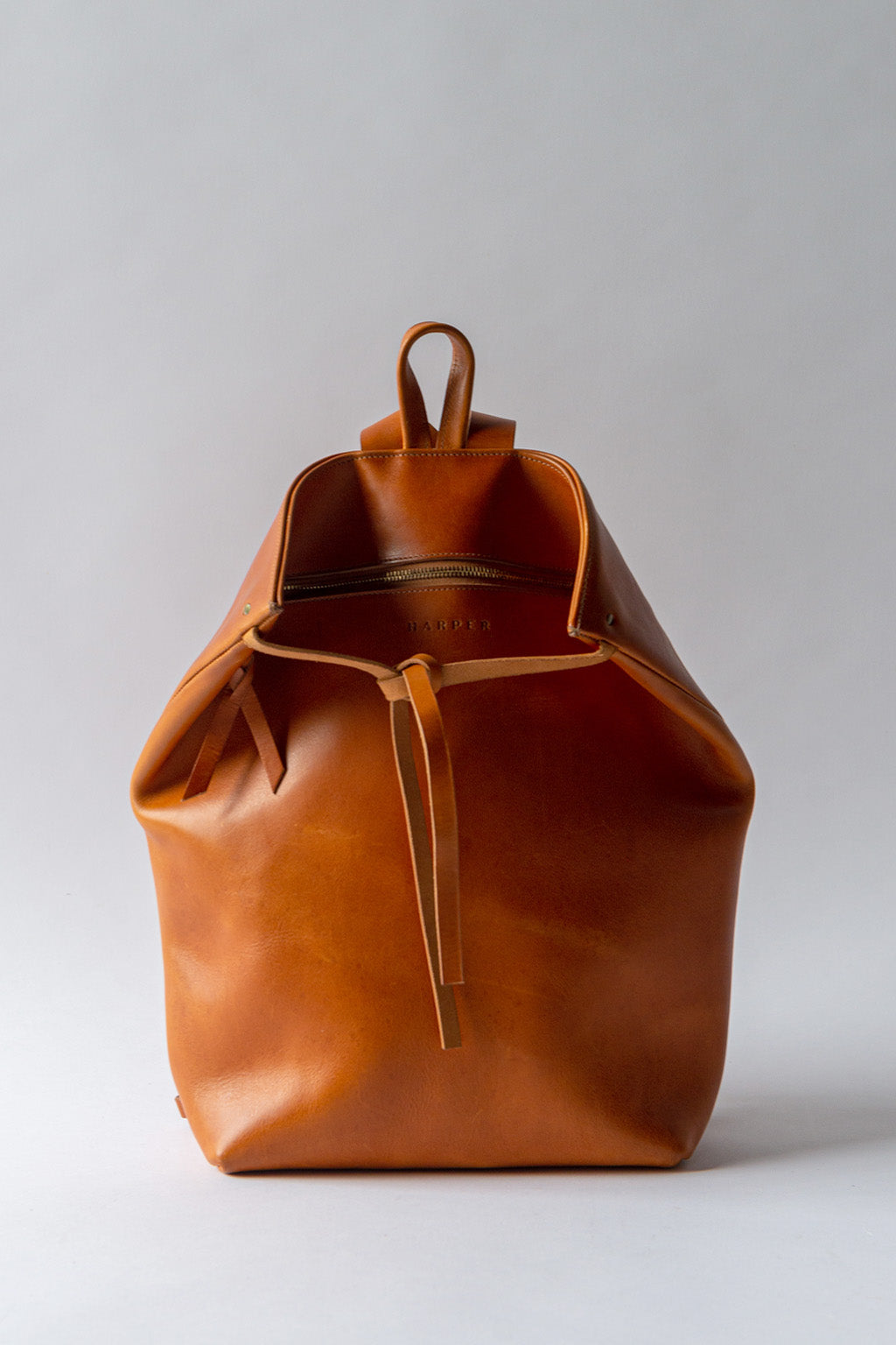A cognac brown leather backpack rests against a white background. The top corners of the backpack are cinched with straps that knot over a zipper, creating a unique shape.