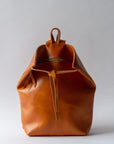 A cognac brown leather backpack rests against a white background. The top corners of the backpack are cinched with straps that knot over a zipper, creating a unique shape.