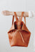 A woman holds out her arm, which is looped through the shoulder straps of a cognac brown leather backpack. The top corners of the backpack are cinched with straps that knot over a zipper, creating a unique shape.
