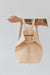A woman holds out her arm, which is looped through the shoulder straps of an undyed leather backpack. The top corners of the backpack are cinched with straps that knot over a zipper, creating a unique shape.