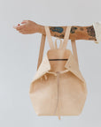 A woman holds out her arm, which is looped through the shoulder straps of an undyed leather backpack. The top corners of the backpack are cinched with straps that knot over a zipper, creating a unique shape.